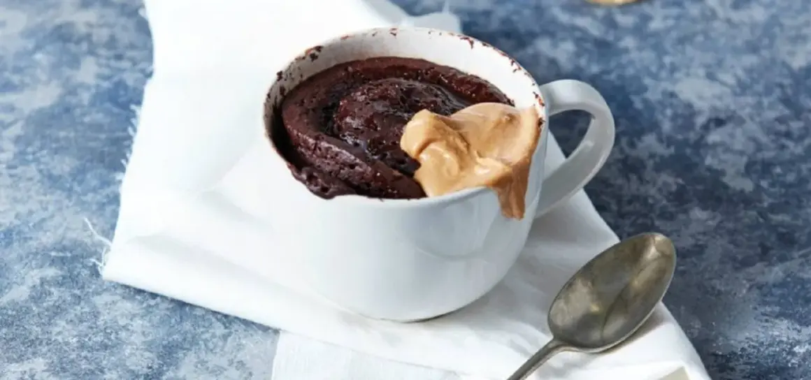 Schokoladiger high protein tassenkuchen mit Erdnussbutter-Topping in einer weißen Tasse, daneben ein Löffel.