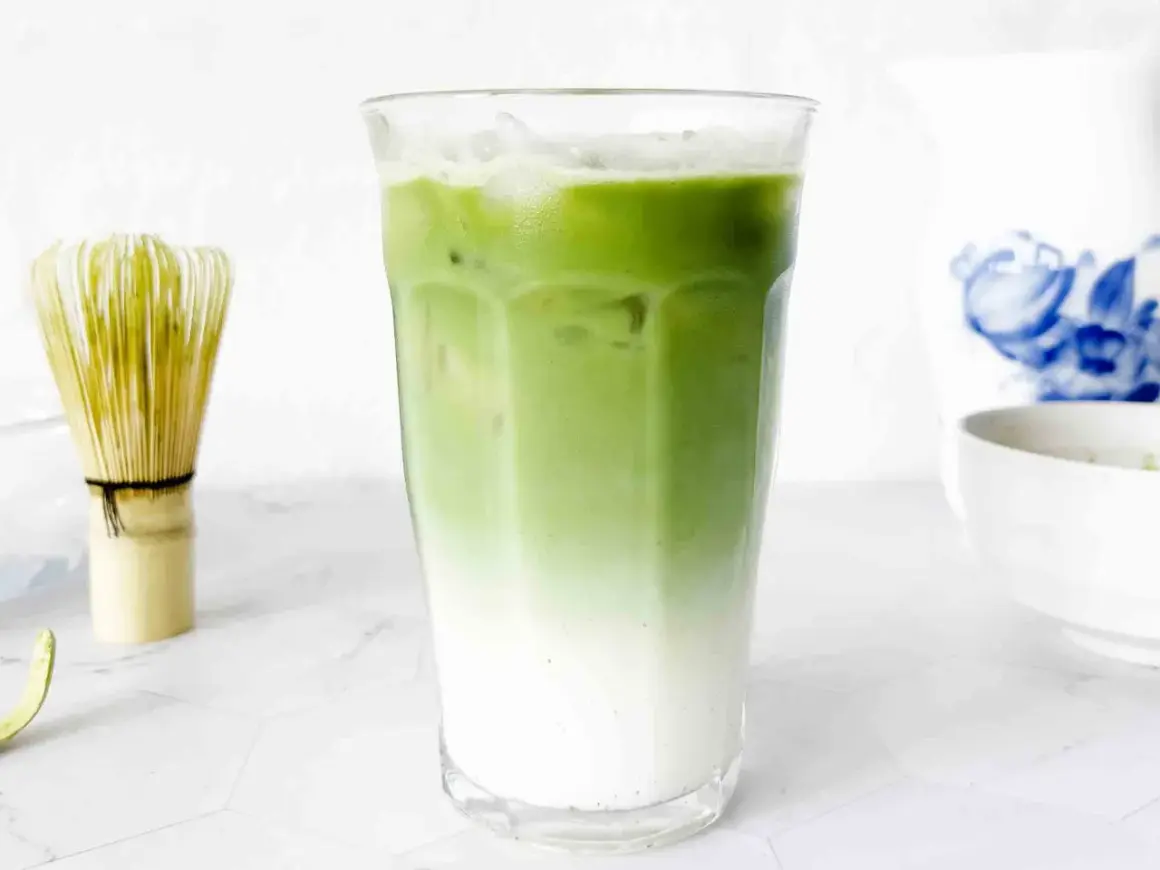 En snygg, isig matcha latte med lager av mjölk och grönt te. Bredvid står en visp för att göra matcha.