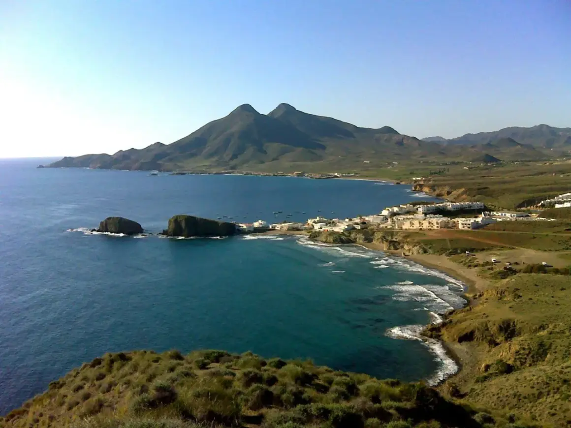 Vista panorámica de la cala del Toro, con sus aguas turquesas, rocas emergiendo y un pueblo blanco al fondo, bajo montañas verdes.