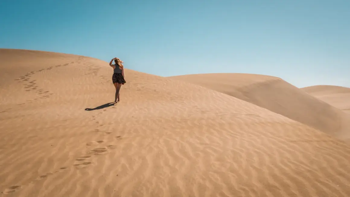 Kobieta spaceruje po piaszczystych wydmach Dunas de Maspalomas, zostawiając ślady na piasku pod błękitnym niebem.