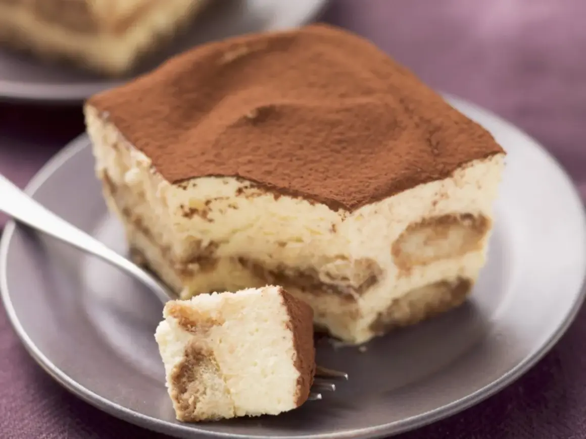 Ein Stück cremiges Tiramisu ohne Zucker, bestäubt mit Kakao, auf einem Teller mit einer Gabel.