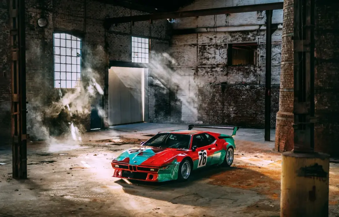 Un m1 coche de carreras con librea roja y verde, número 76, descansa en un almacén polvoriento con rayos de sol y humo.