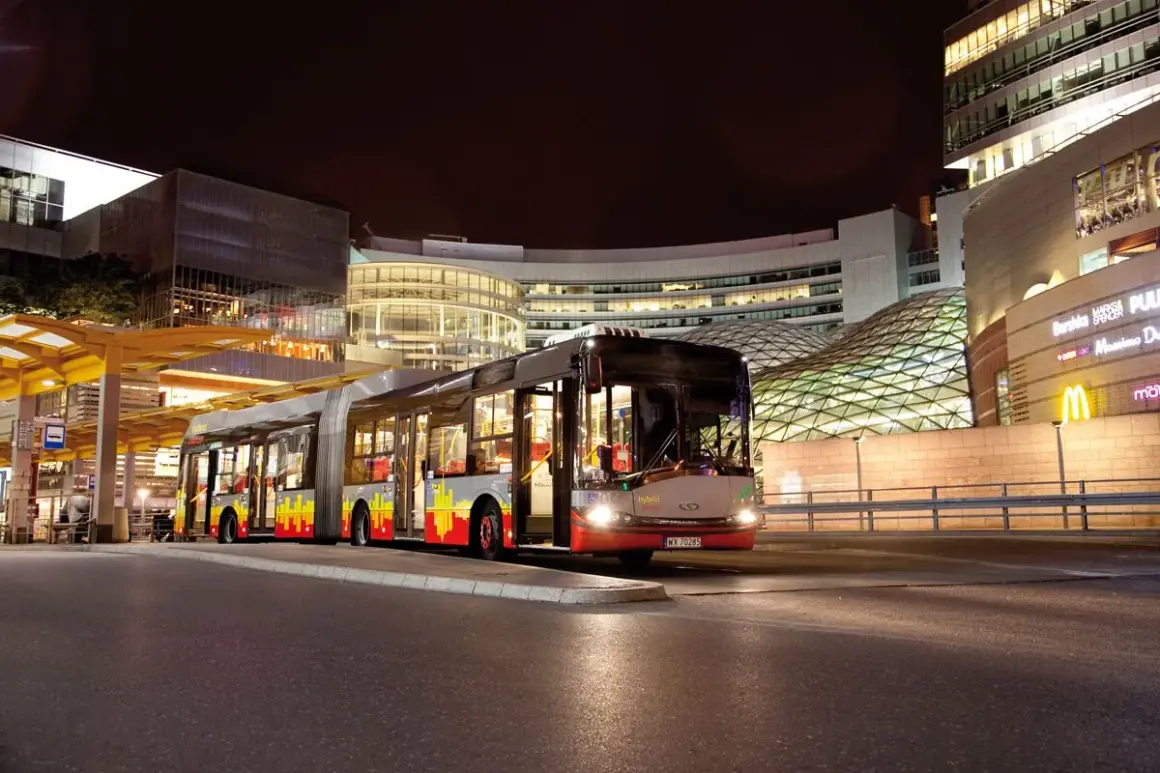 Długi, przegubowy autobus linii 59 w Łodzi nocą, zatrzymuje się przy nowoczesnym centrum handlowym.