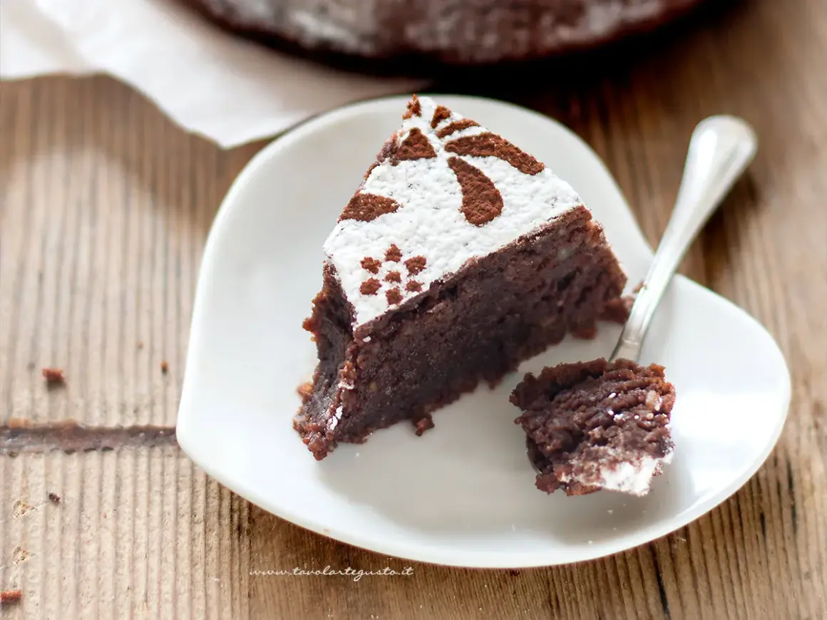Una fetta di torta caprese ricetta originale napoletana, cosparsa di zucchero a velo e cacao, su un piatto bianco a forma di cuore.