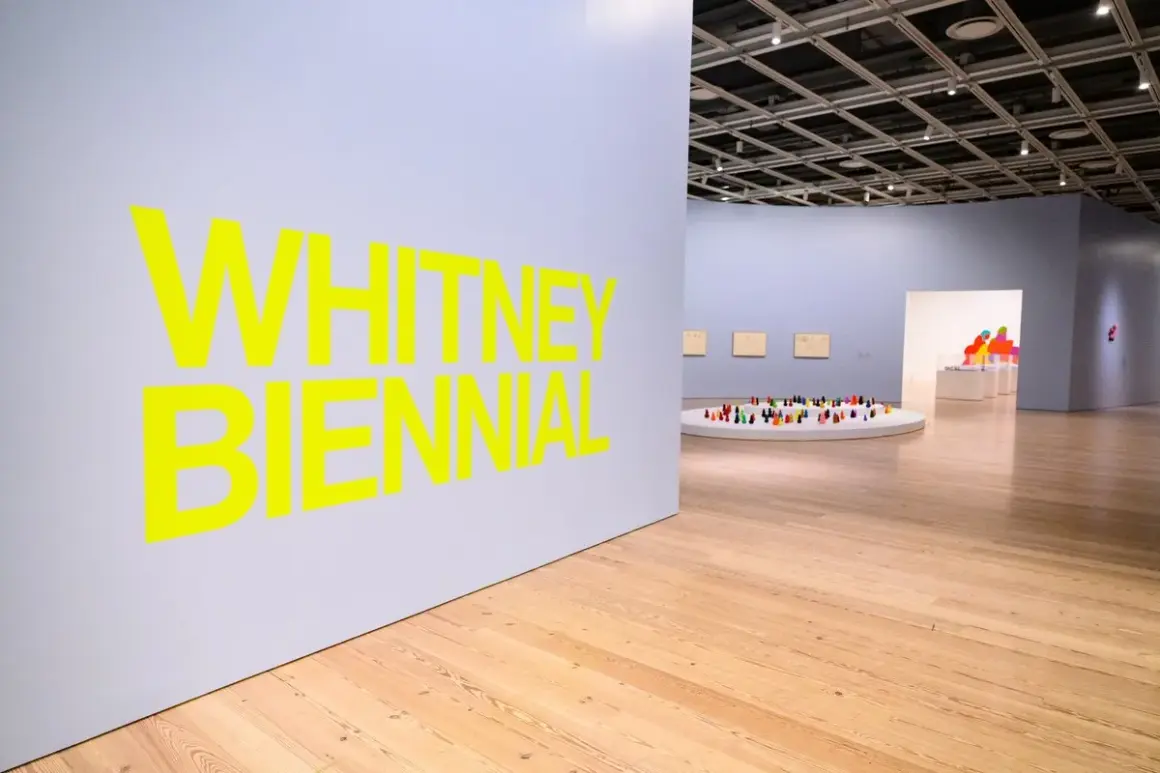 Bright yellow text announces the Whitney Biennial 2026. Inside the gallery, a circular display holds a colorful collection of small figures.
