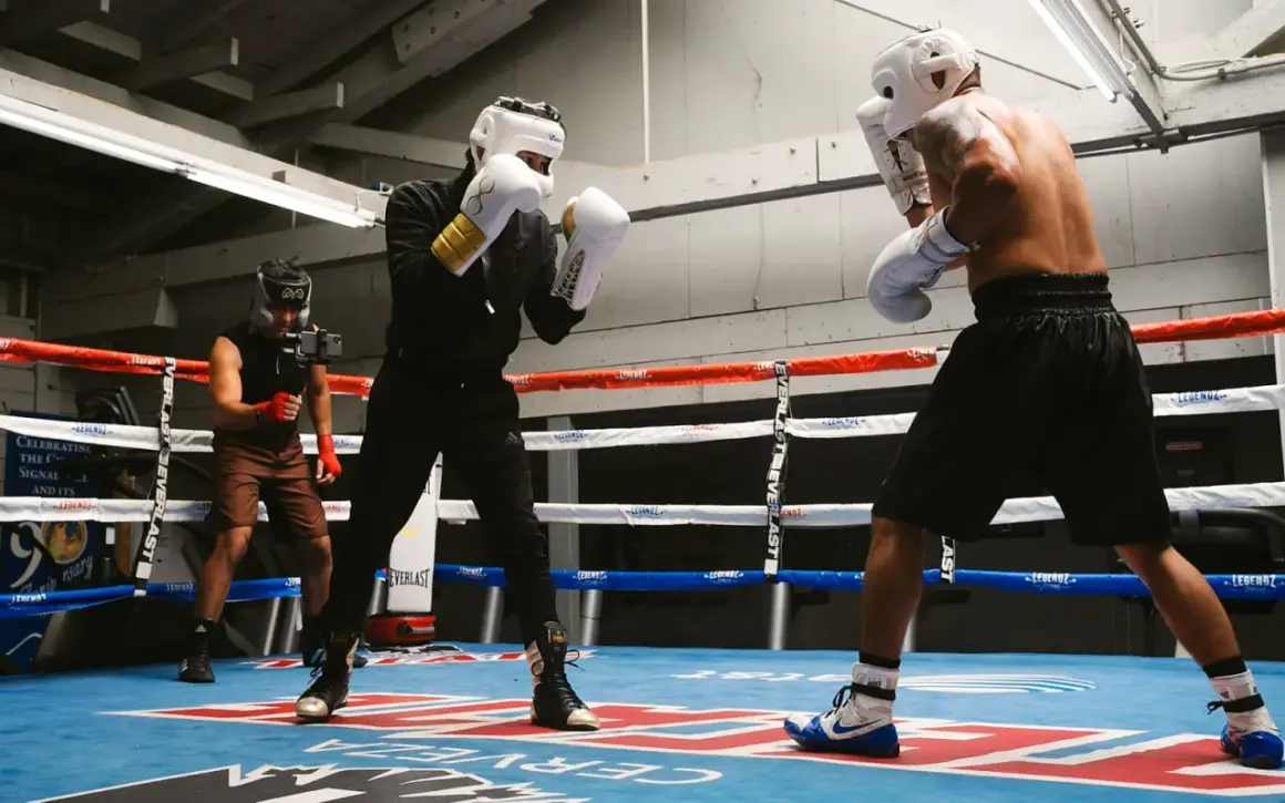 Two boxers in a brawler boxing style spar in a gym, one with white gloves and gold accents, the other shirtless. A third person films them.