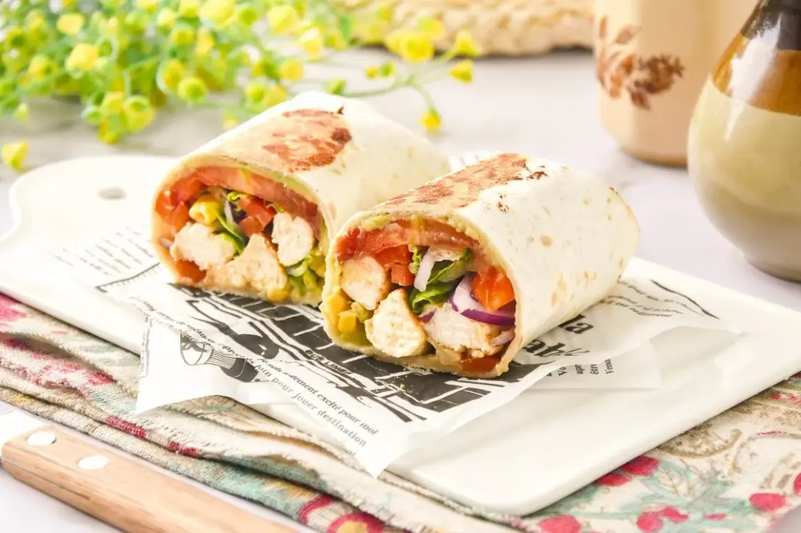 Dos burritos de pollo cortados por la mitad, rellenos de pollo, tomate, lechuga y cebolla morada, servidos sobre papel de periódico.