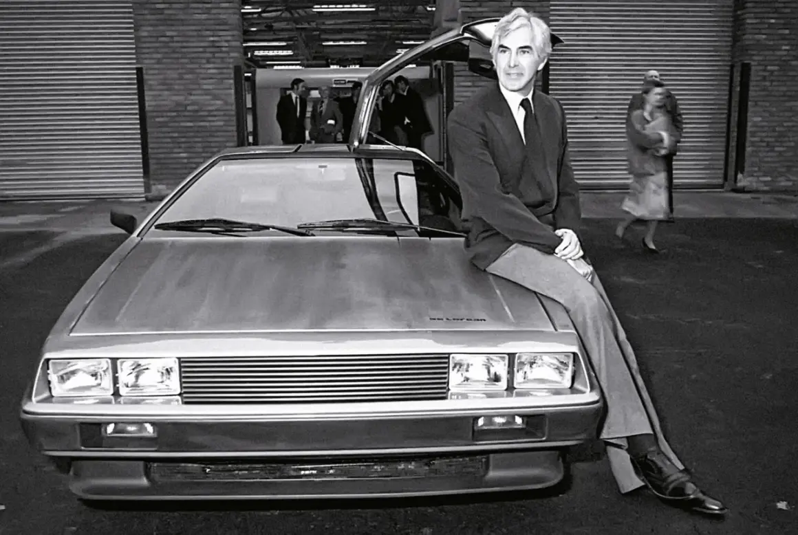 Mężczyzna w garniturze siedzi na masce kultowego **DeLorean**a z otwartymi drzwiami. W tle widać ludzi i budynek.