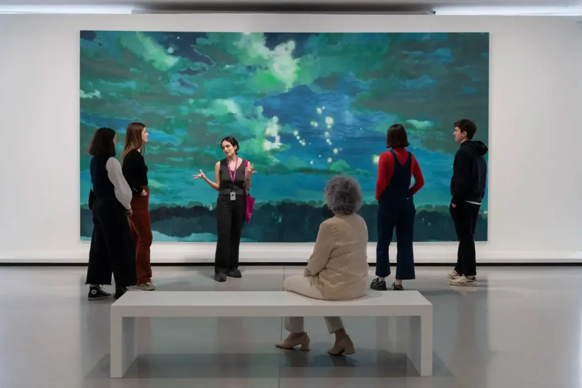 Visite guidée d'une œuvre d'art abstraite aux teintes bleues et vertes, évoquant une scène céleste. Un groupe écoute attentivement, comme à la bourse du commerce.