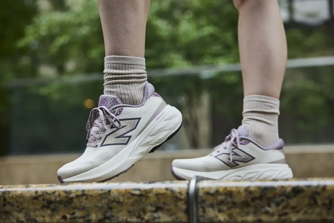 Scarpe da tennis New Balance in beige e viola, con calzini beige arricciati, su uno sfondo verde sfocato.