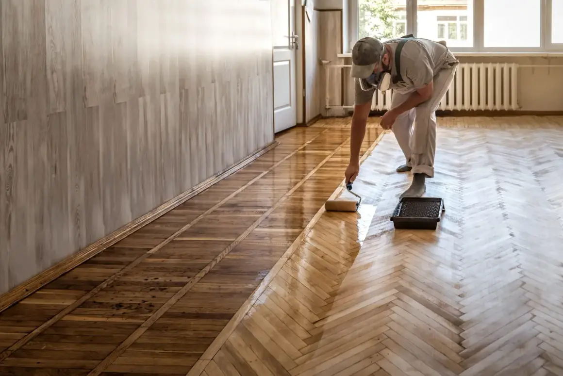 Un homme, masqué, utilise un rouleau pour vitrifier ou cirer un parquet en bois, lui redonnant son éclat.