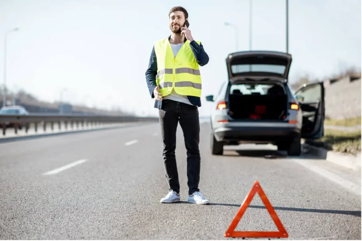 Mann mit Warnweste telefoniert neben seinem Auto mit offenem Kofferraum. Ein Warndreieck liegt auf der Straße. Wie viele Warnwesten im Auto sind, ist unklar.