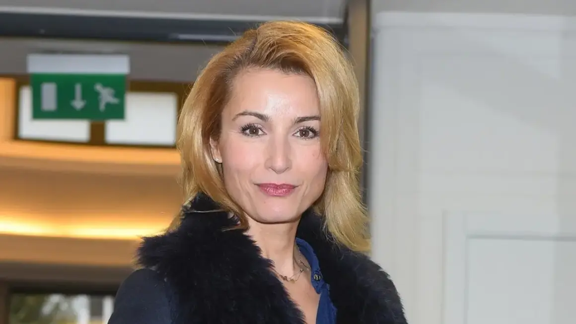 Joanna Brodzik uśmiecha się, ubrana w czarny płaszcz z futrzanym kołnierzem. W tle zielony znak ewakuacyjny.