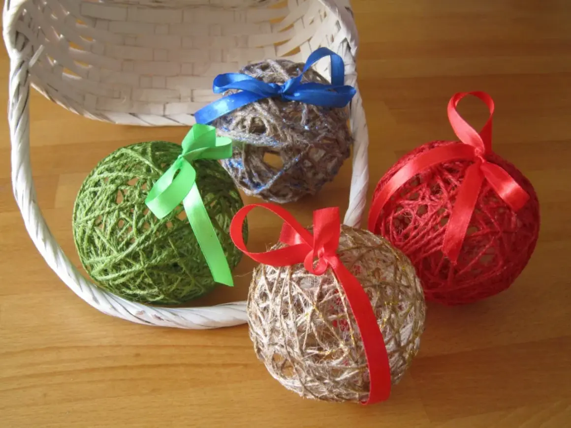 Palline decorative fatte con spago e colla vinilica, decorate con nastri colorati, appoggiate vicino a un cestino bianco.