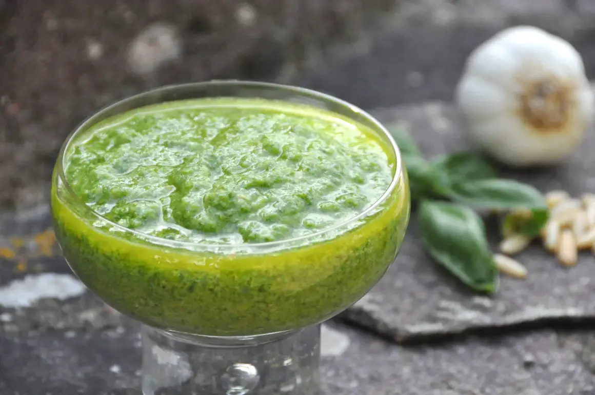 Pesto di basilico bimby in una coppetta, con aglio, pinoli e foglie di basilico sullo sfondo.