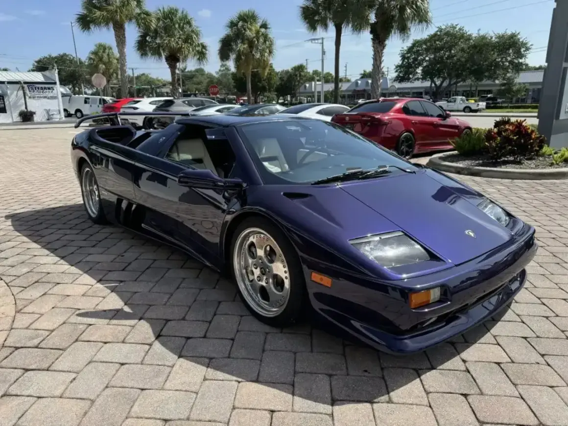 Fioletowy Lamborghini Diablo z jasnym wnętrzem, zaparkowany na brukowanym podjeździe, z palmami w tle.