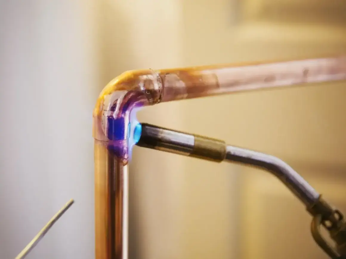 Un chalumeau chauffe un raccord coudé en cuivre pour le souder. La flamme bleue et violette illumine le métal.
