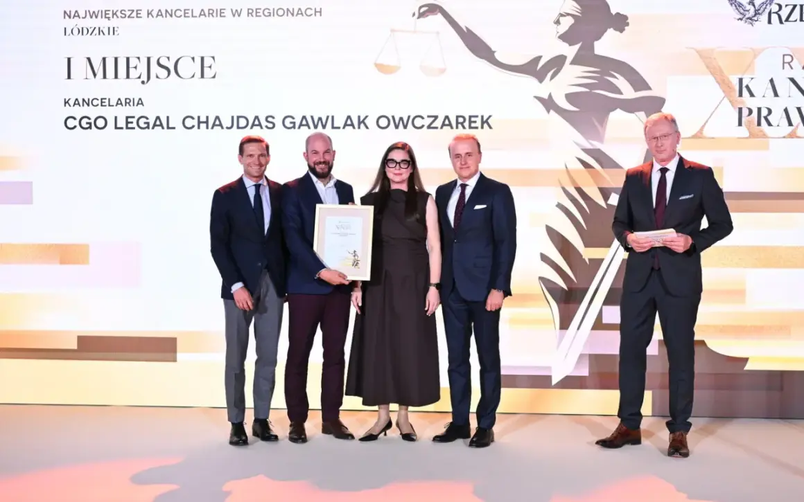 Cztery osoby w garniturach i sukience stoją na scenie, odbierając nagrodę.