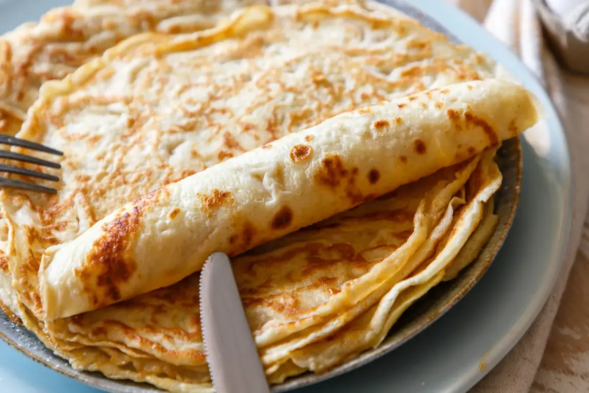 Ein Stapel goldbrauner Eierkuchen, einer davon aufgerollt, liegt auf einem Teller. Ein Messer und eine Gabel liegen daneben. Perfekt für ein einfaches Eierkuchen Rezept.