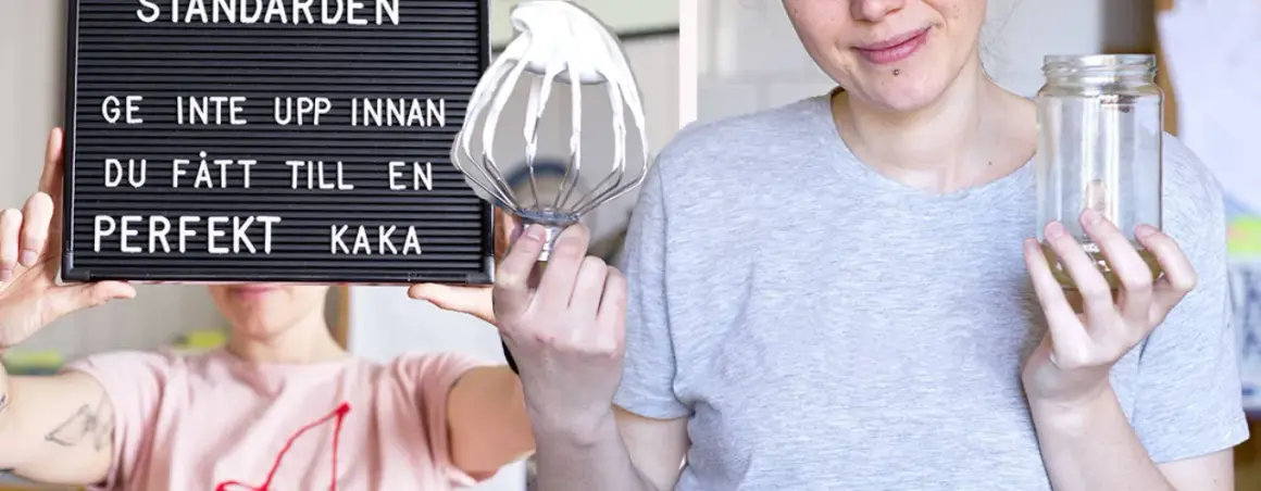 En person håller upp en skylt med texten "Ge inte upp innan du fått till en perfekt kaka". Bredvid syns en visp med grädde och en tom burk, redo för en vegansk fika.