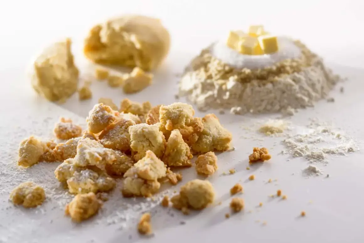 Knusprige Streusel mit gemahlenen Mandeln, bestäubt mit Puderzucker. Im Hintergrund Teig und Zutaten für eine köstliche Leckerei.