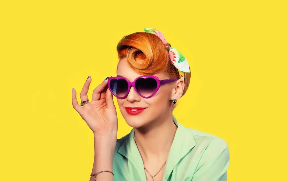 Une femme au style rockabilly, cheveux coiffés en chignon, porte des lunettes violettes en forme de cœur et un foulard coloré.