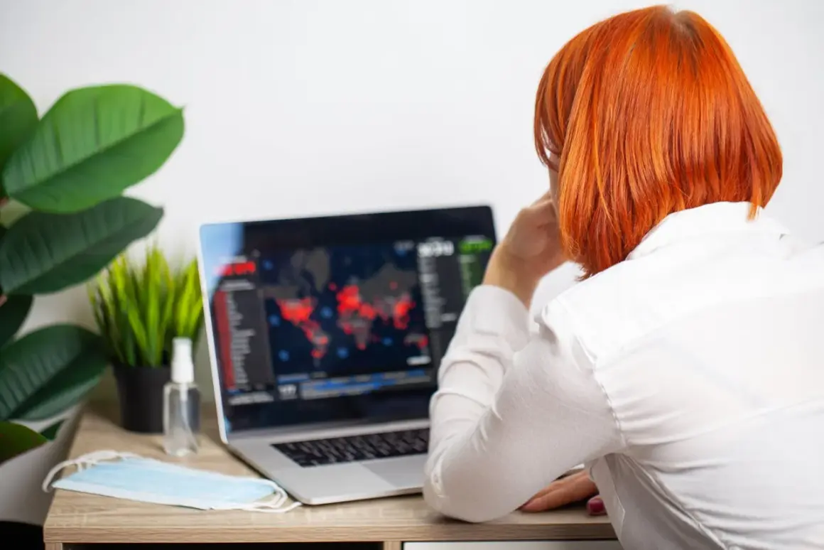 Kobieta z rudymi włosami patrzy na ekran laptopa z mapą świata i danymi. Obok leży maseczka i płyn do dezynfekcji.