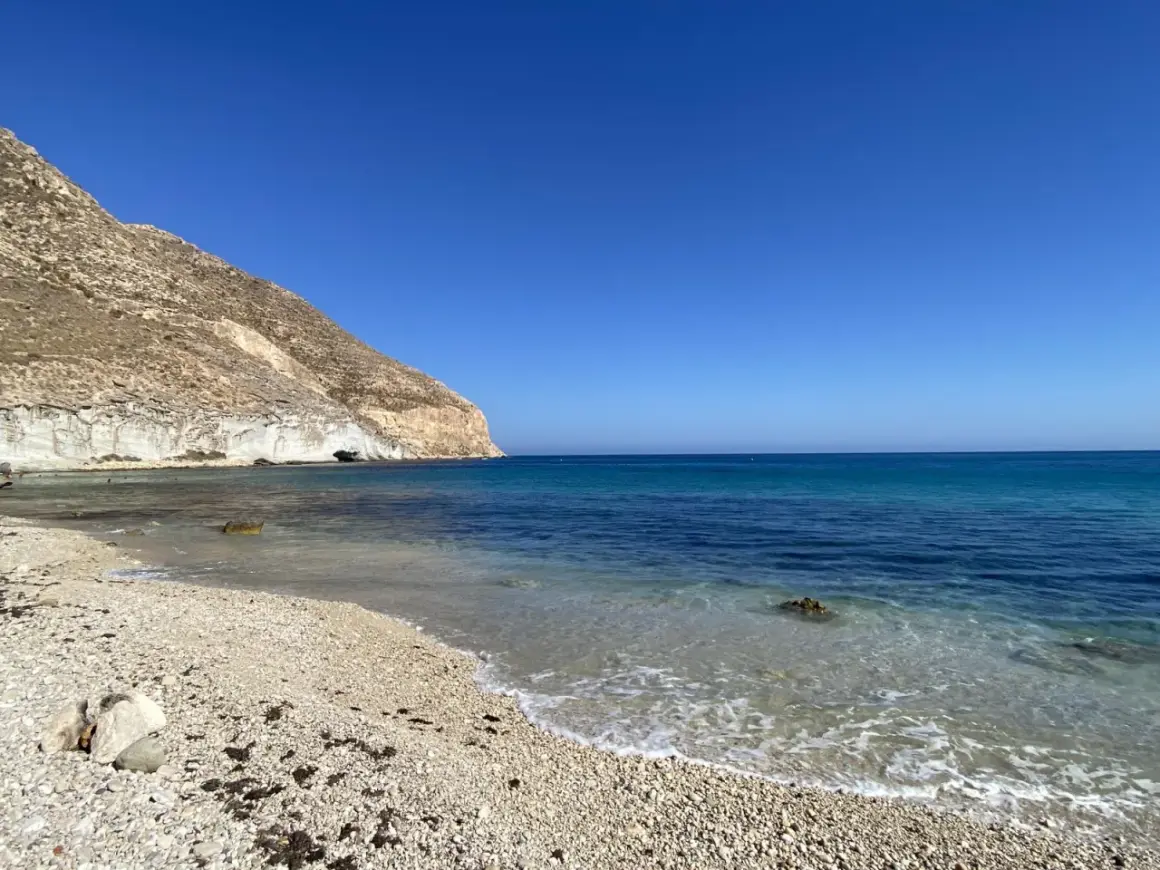 La **playa Peñón Blanco** se extiende bajo un cielo azul intenso. Las olas acarician la orilla de guijarros, mientras una imponente montaña rocosa se alza a la izquierda, con una cueva marina visible en su base.