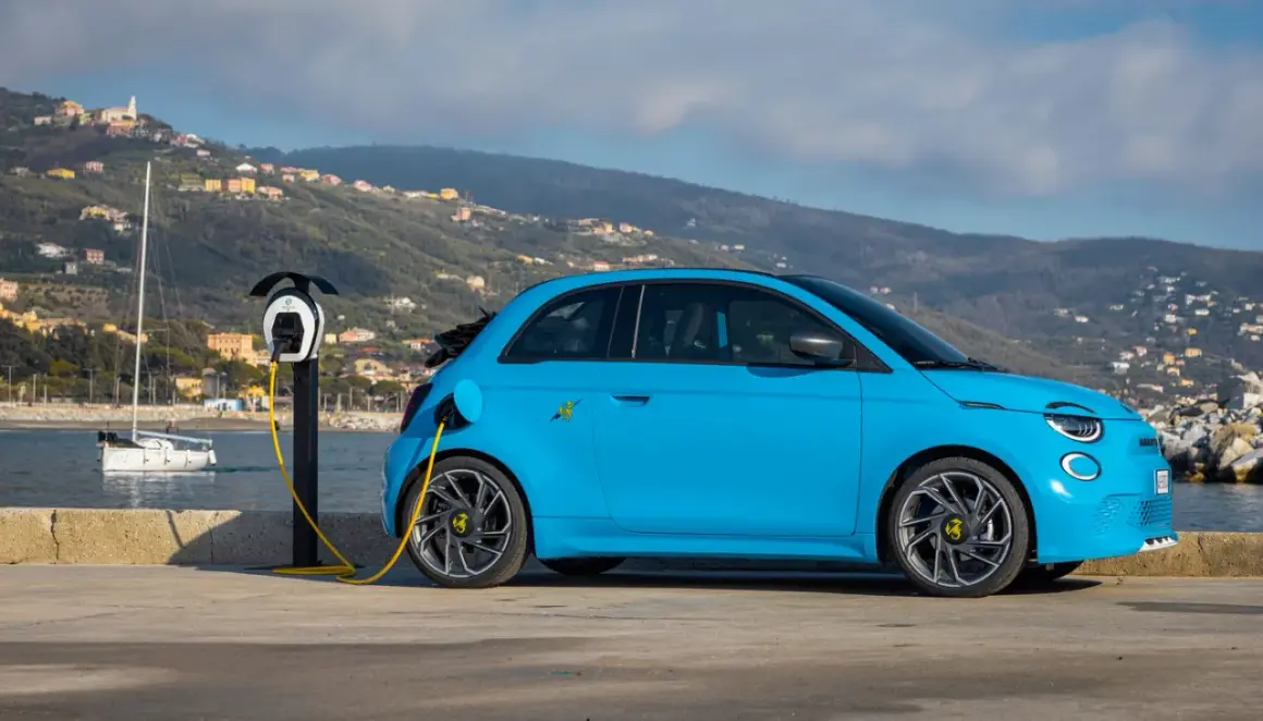 Abarth EV azul cargando junto al mar, con un velero y colinas al fondo.