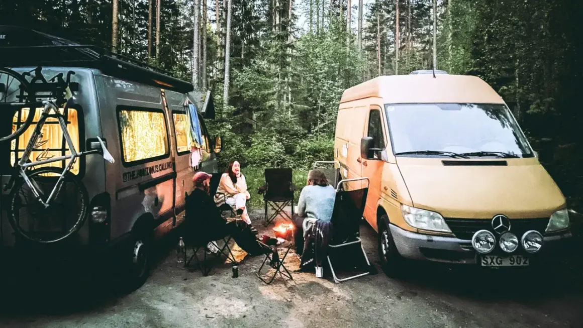 Två husbilar parkerade i skogen, med folk som sitter runt en lägereld. En cykel sitter på takräcket på den ena bilen. En härlig bild av van life.