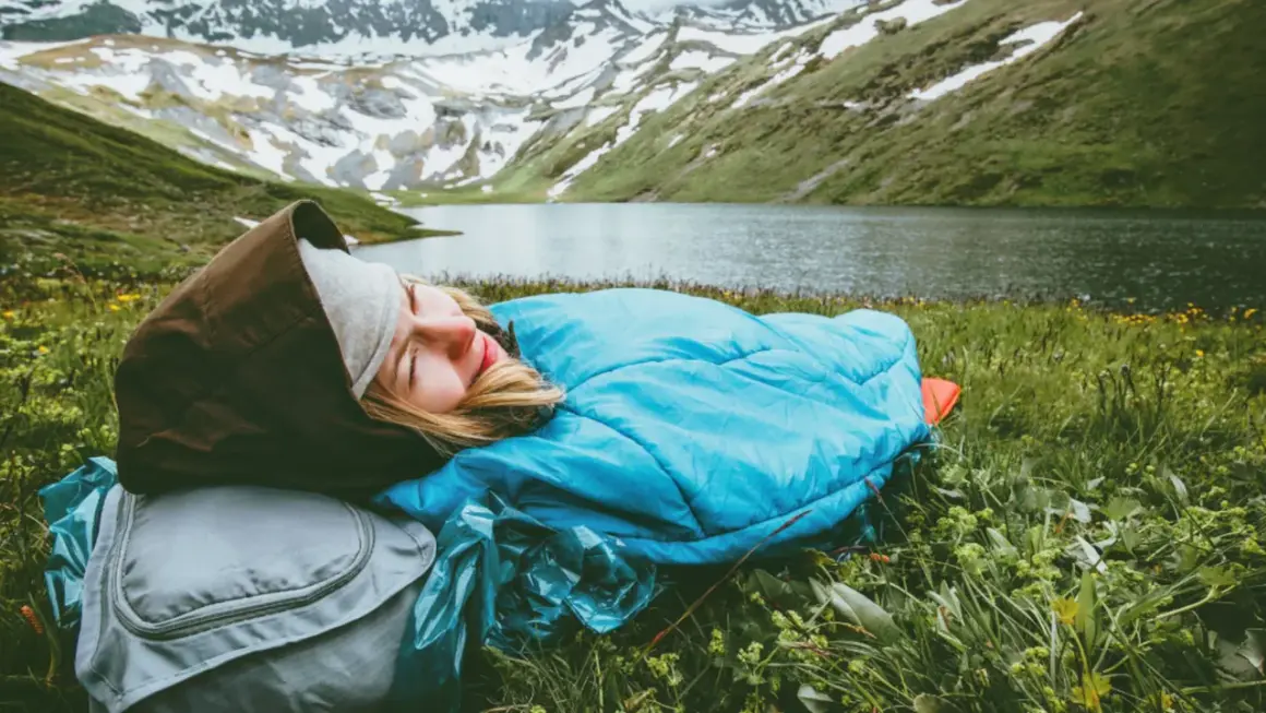 Persona durmiendo en saco azul, con mochila gris, en un prado verde junto a un lago y montañas nevadas. Vivac acampada perfecta.