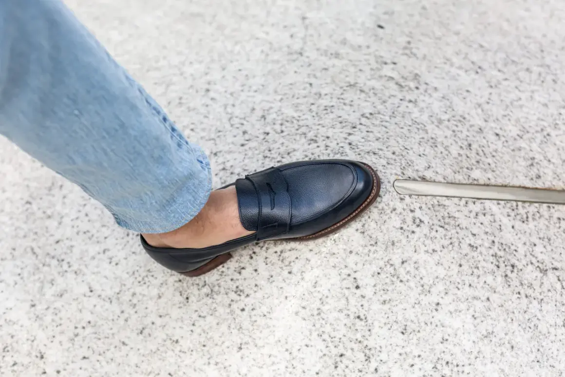 Un homme en jean bleu porte une chaussure de ville marine. L'outil à côté suggère une envie de ressemeler chaussures.