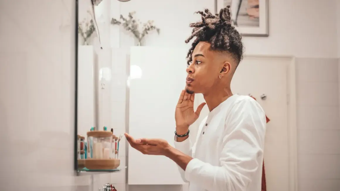 Mann mit Dreadlocks im Spiegel, der sich die Haut richtig reinigen möchte.