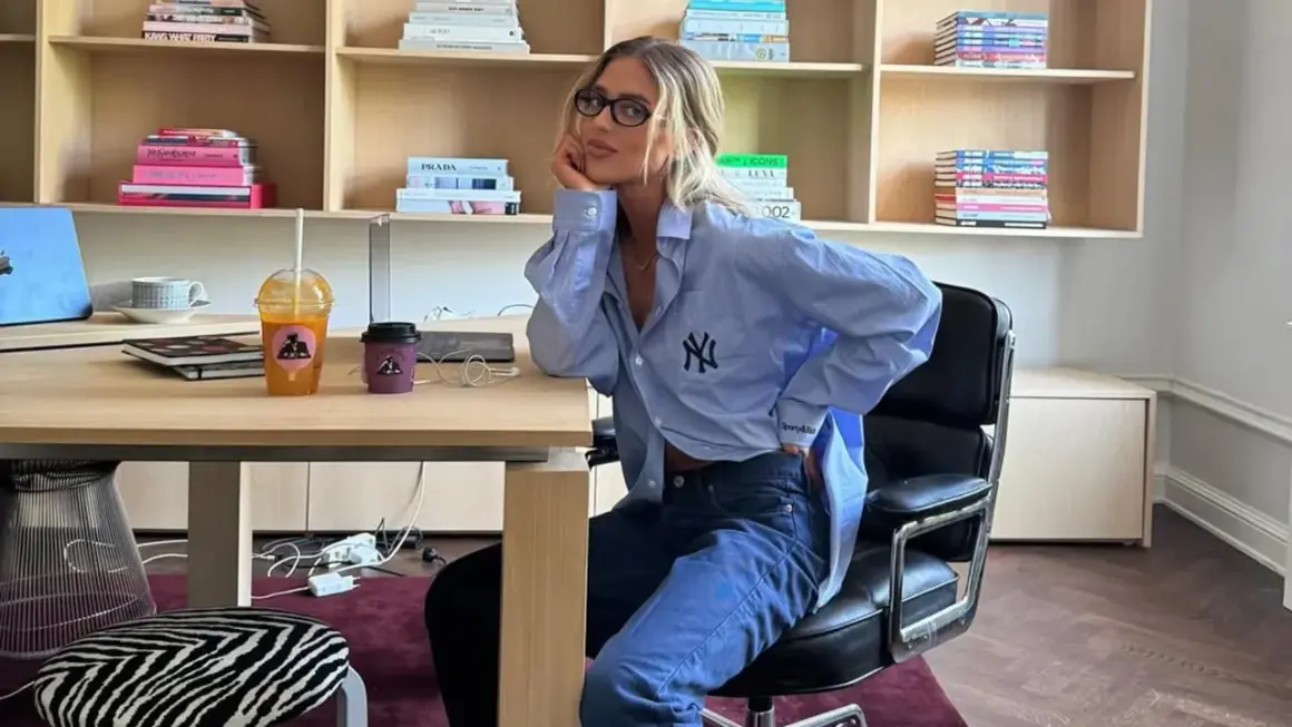 Chica con gafas sentada en una silla de oficina, luciendo un outfit oficina verano hombre: camisa azul oversize y vaqueros.