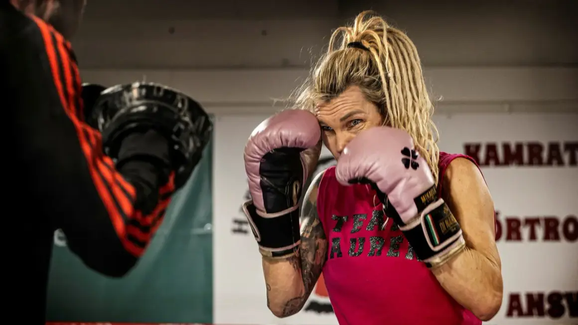 Mikaela boxare i rosa handskar, redo för träning. Hennes blick är fokuserad, med dreadlocks som ramar in ansiktet.