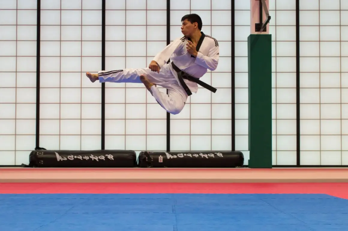 Man i vit dräkt med svart bälte utför ett högt spark i luften under en tae kwon do-träning.