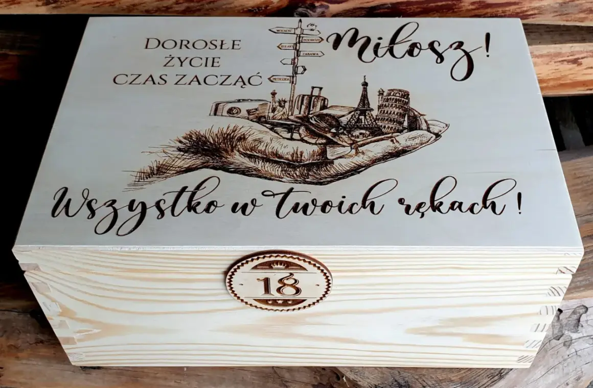 Drewniana skrzynka z grawerem na 18 urodziny od chrzestnej. Na wieczku napis "Dorosłe życie czas zacząć, Miłosz! Wszystko w Twoich rękach!" i grafika dłoni z symbolami podróży.