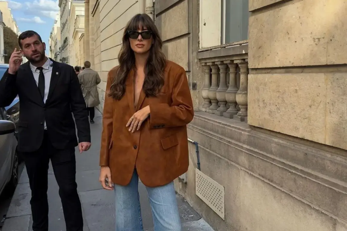 En kvinna bär en snygg, brun vero moda mockajacka och jeans på en stadsgata. En livvakt går bakom henne.