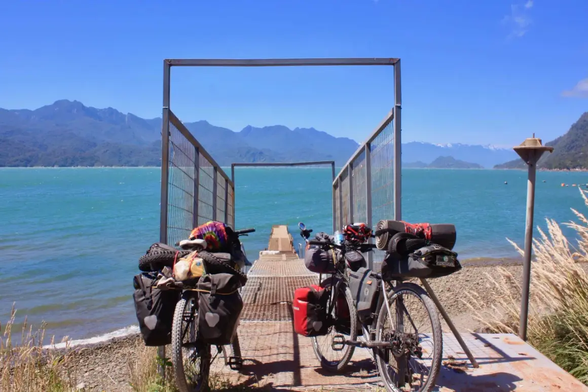 Dos bicicletas equipadas para aventura esperan junto a un muelle en la Carretera Austral. El agua turquesa y las montañas azules crean un paisaje impresionante.