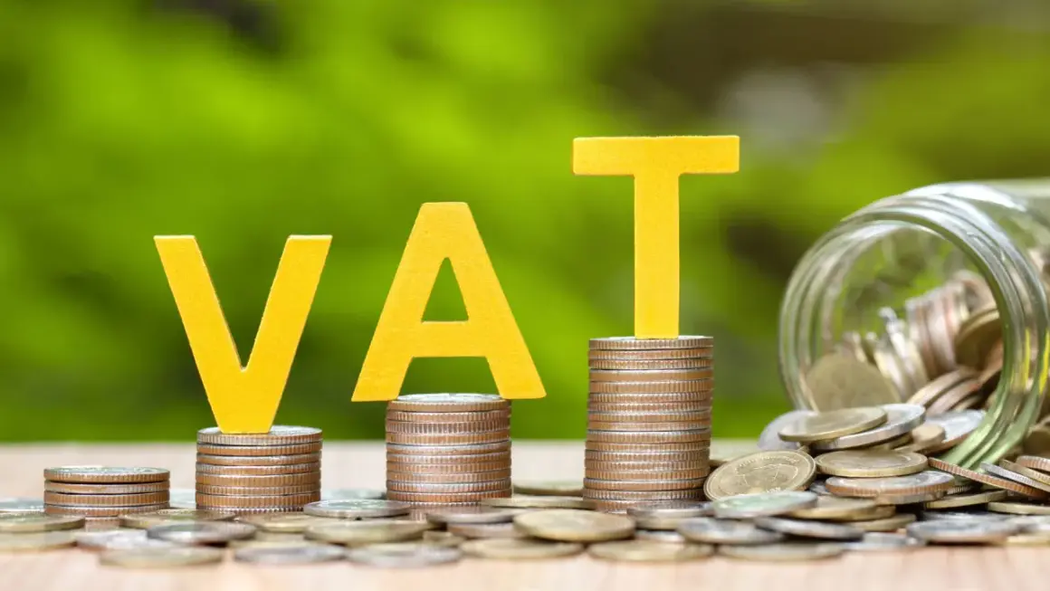 Żółte litery VAT na stosach monet, obok słoik pełen pieniędzy. Grupa vat to ważny element finansów.