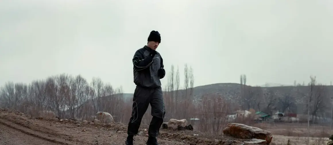 A boxer in a black tracksuit and beanie is running outdoors, practicing his footwork. The barren landscape suggests a cold day for this intense training.