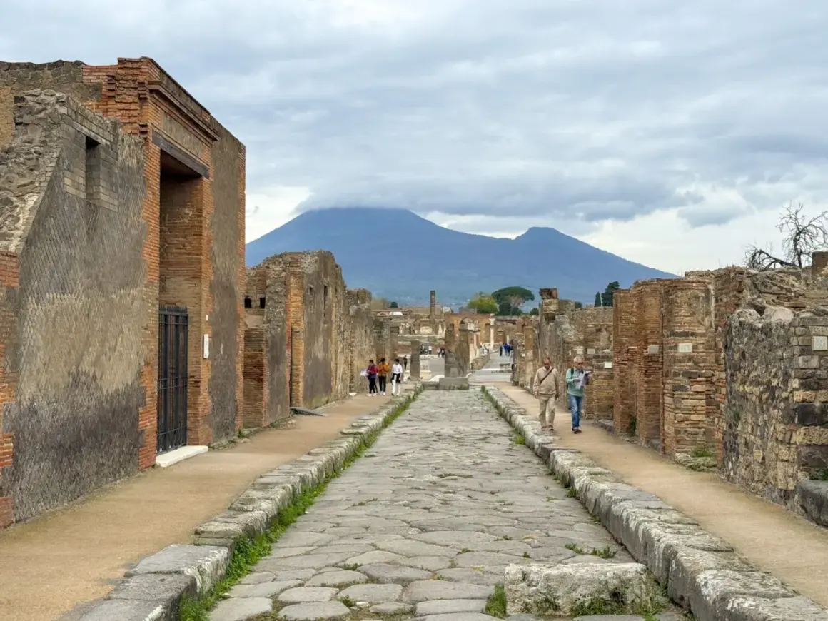 Antigua calle empedrada en el parque arqueológico de Pompeya, con ruinas a ambos lados y el Vesubio al fondo.