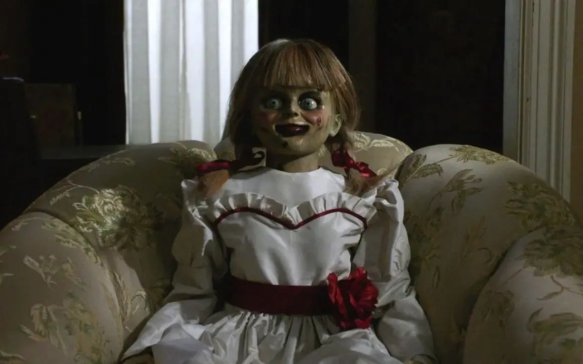 Den läskiga riktiga Annabelle dockan sitter i en blommig soffa, med sina stora ögon och röda rosett.