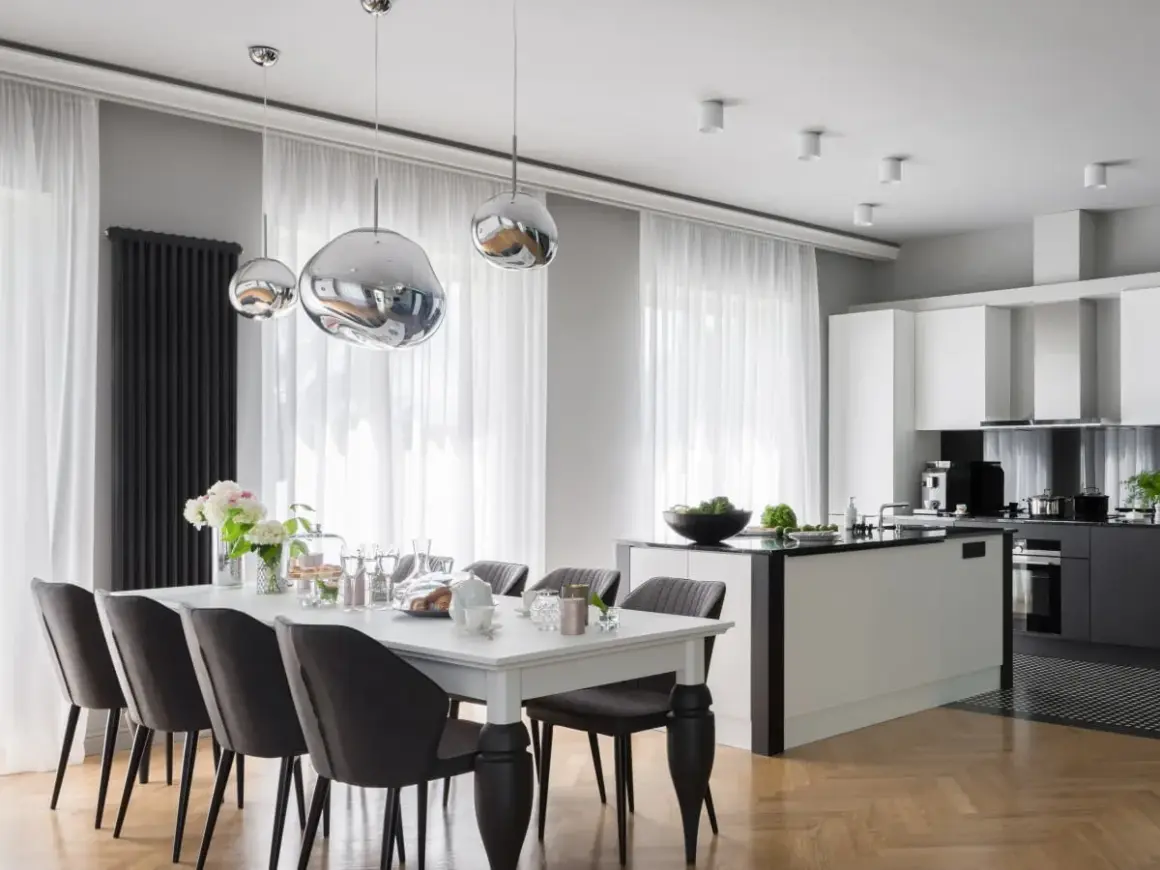 Elegancki styl glamour salon z kuchnią. Nowoczesne lampy, biała wyspa kuchenna i stół z szarymi krzesłami tworzą spójną całość.