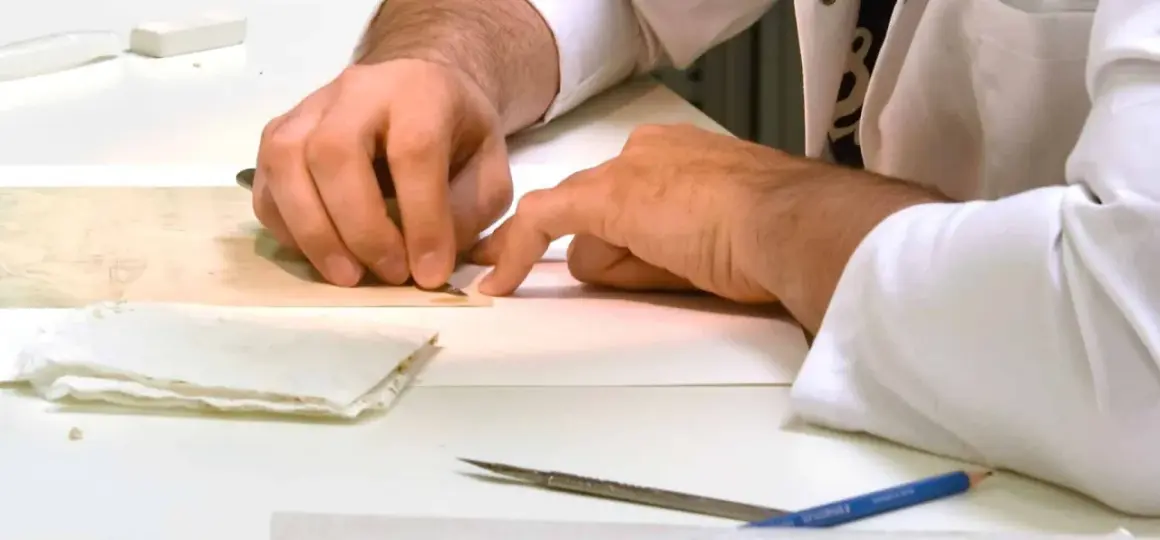 Main tenant avec précision un scalpel pour enlever de la colle sur un document ancien. Des outils et du papier sont disposés sur la table.