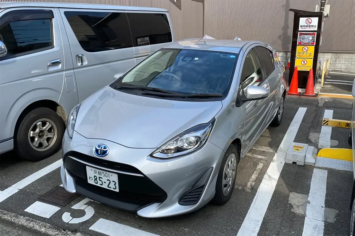 Srebrny samochód na parkingu w Japonii, gdzie obowiązuje ruch lewostronny. Obok stoi srebrny van.