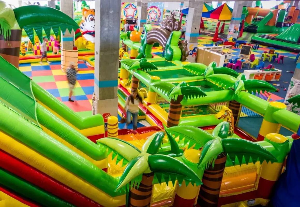 Kolorowy park rozrywki we Wrocławiu z dmuchanymi zjeżdżalniami i palmami. Dzieci biegają po kolorowej macie.