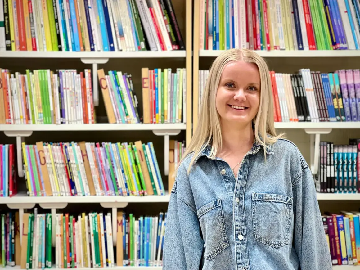 Evelina Persson ler glatt framför bokhyllor fyllda med färgglada böcker.