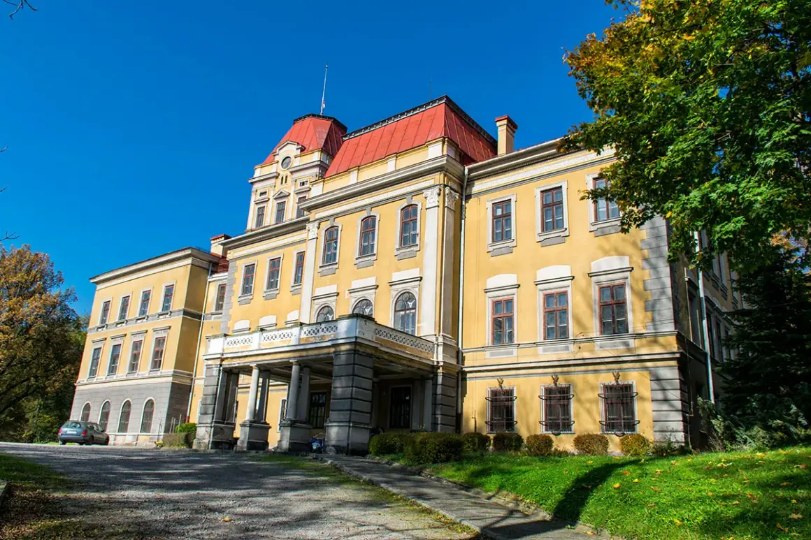 Żółty pałac z czerwonym dachem, otoczony zielenią i jesiennymi drzewami. W oddali widać kończyce wielkie.
