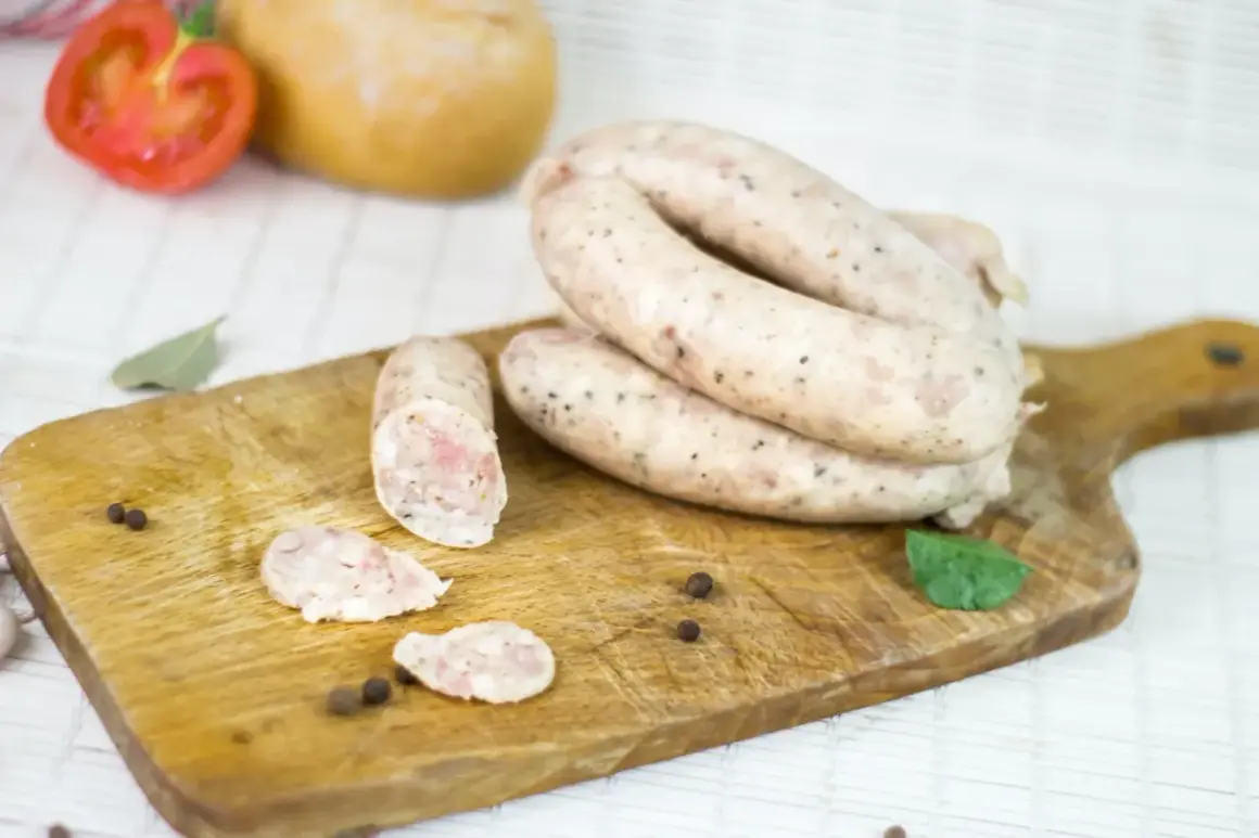 Biała kiełbasa na desce, obok pomidor, chleb i przyprawy. Zastanawiasz się, ile m mają jelita?